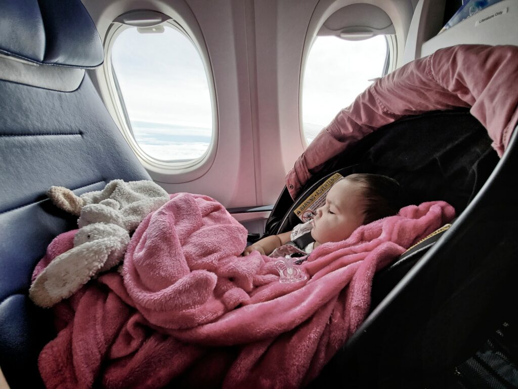 A baby girl is seen sleeping in her car seat while traveling on a plane wrapped in a pink blanket.